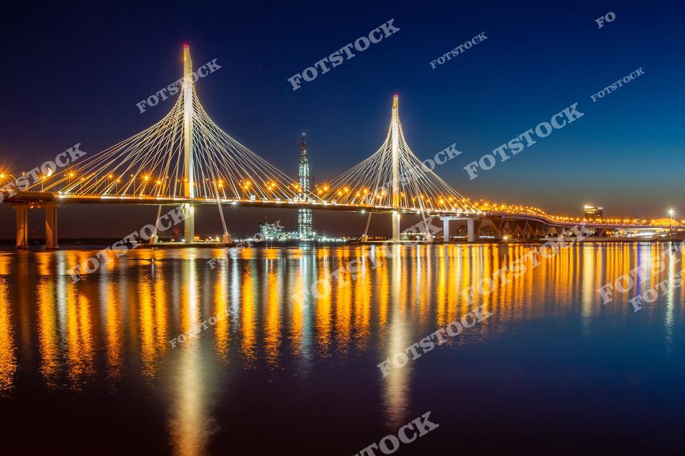 На фотографии изображён Вантовый мост Западного скоростного диаметра (ЗСД) с его эффектной ночной подсветкой. Вдали виднеется небоскрёб. Золотистые огни моста красиво отражаются в водах Финского залива. Этот вид подчеркивает современность и величие Санкт-Петербурга, соединяя элементы транспорта и архитектурных достижений.